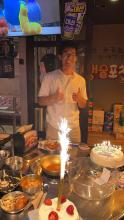 Daniel Ha stands behind a table that holds a cake with a sparkler on top