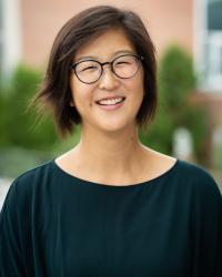 Yui Hashimoto smiling with short dark brown hair and wearing a dark green top, and black round glasses