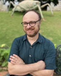 Prof. Joseph Getzoff sitting in a park for a headshot. There is a large grasshopper behind him. 
