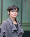 Latiné fem with hair in bun. She wears glasses and a grey sweater over a black shirt. The background is blurred.